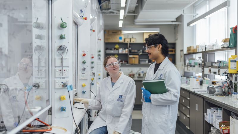 Two students wearing PPE chat to each other in chemistry laboratory 