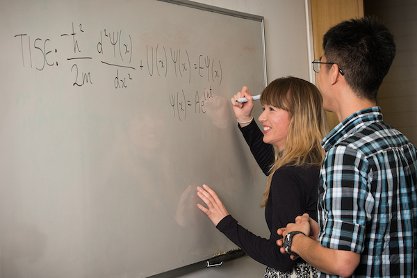A student writing a complex mathematical formula on a whiteboard, while another student looks on
